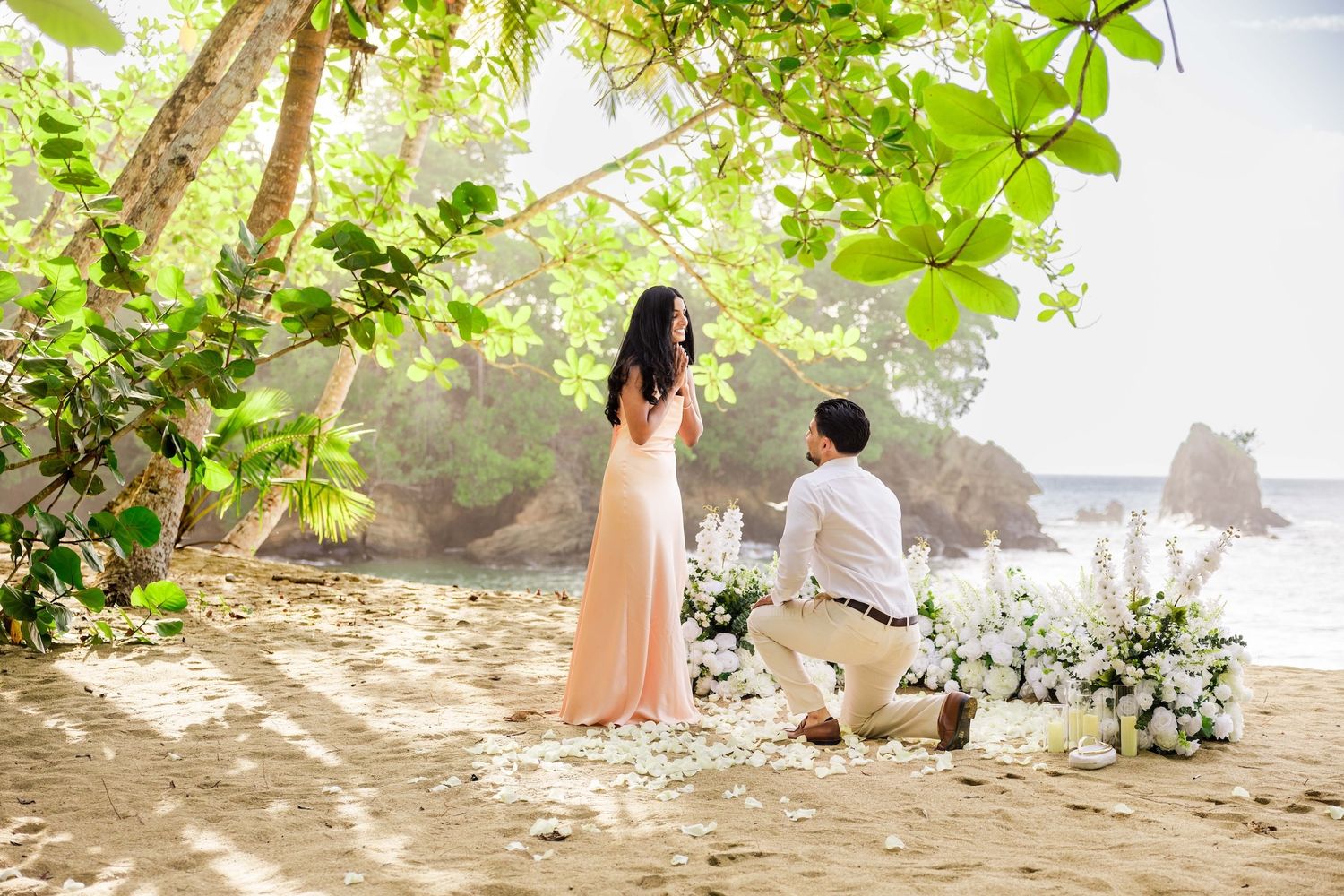 Man kneeling on a tropical beach proposing to a woman in a pink dress surrounded by white flowers.