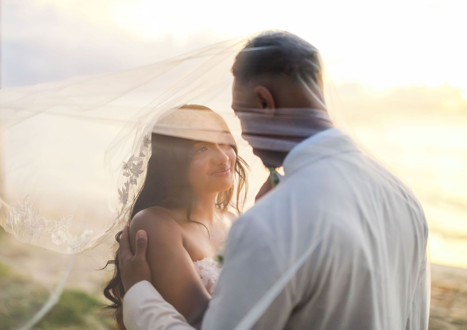 A couple sharing a romantic moment under a flowing wedding veil on a beach at golden sunset.