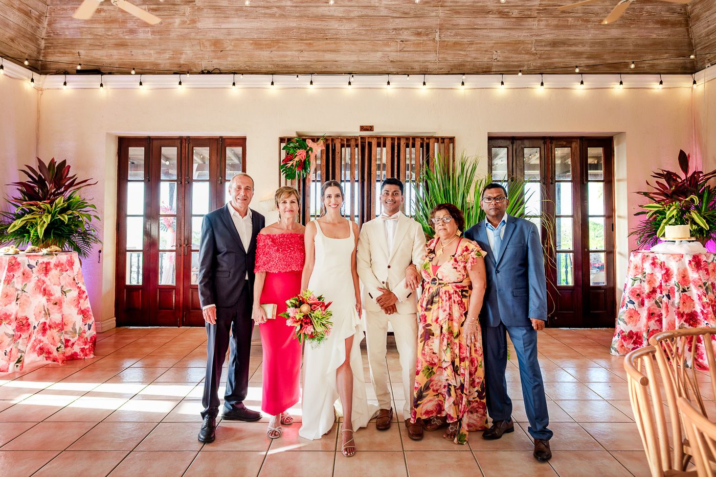Wedding group portrait in formal attire standing in front of wooden French doors with tropical decor.