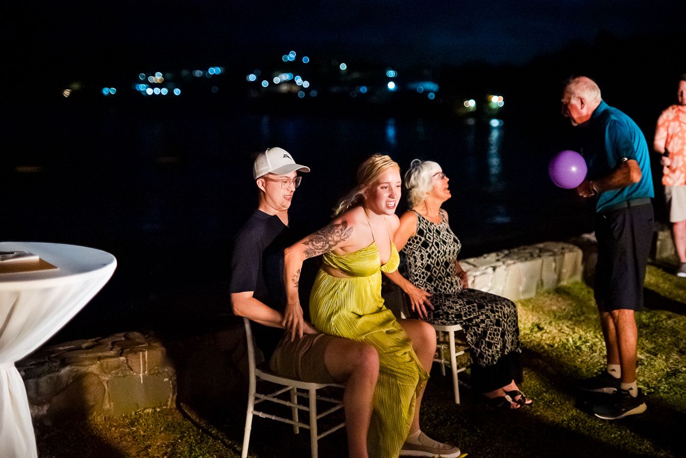 Nighttime gathering on a waterfront patio with city lights twinkling in the background.