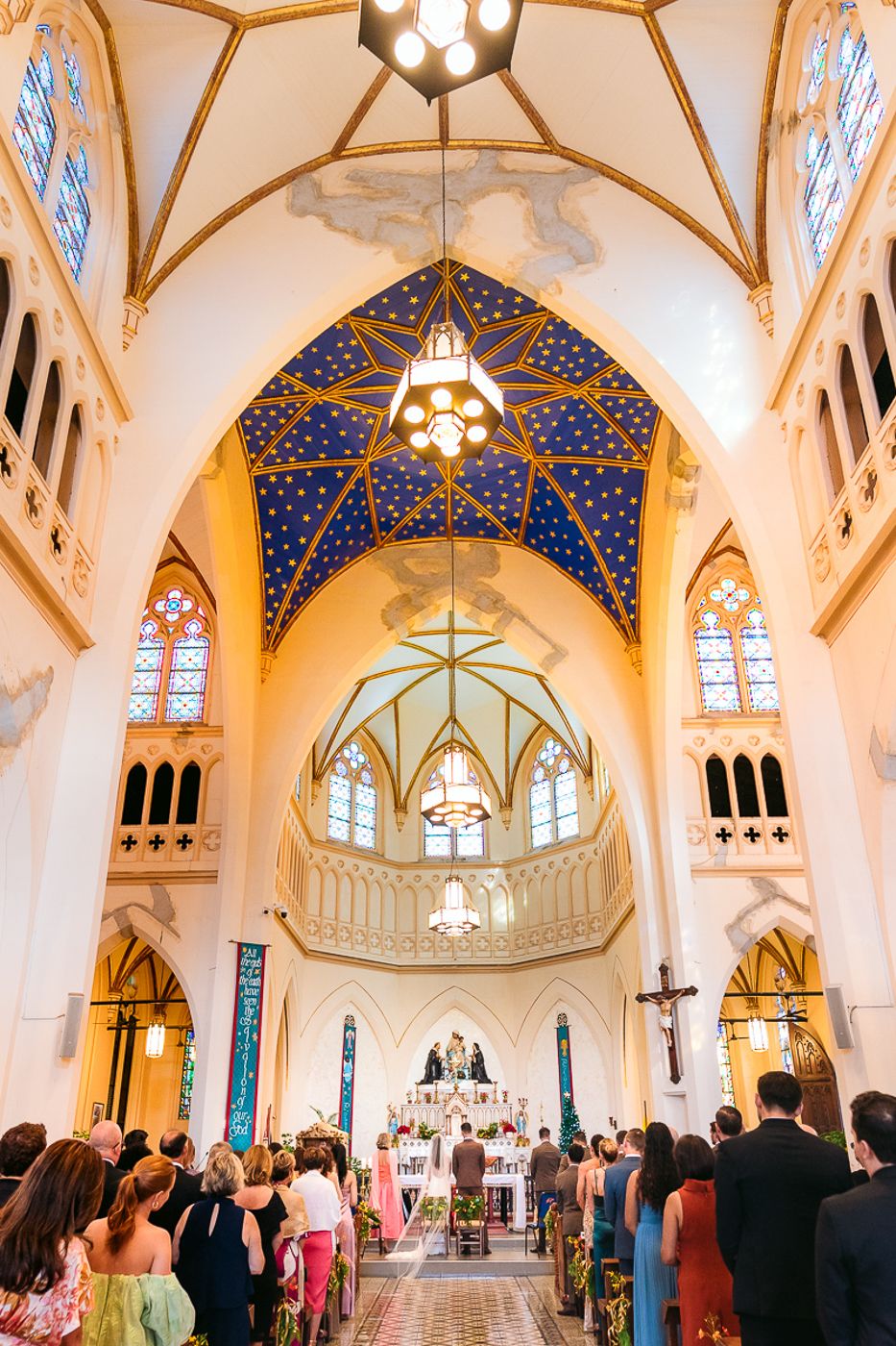 Gothic church interior with vaulted blue starry ceiling and ornate stained glass windows during ceremony.