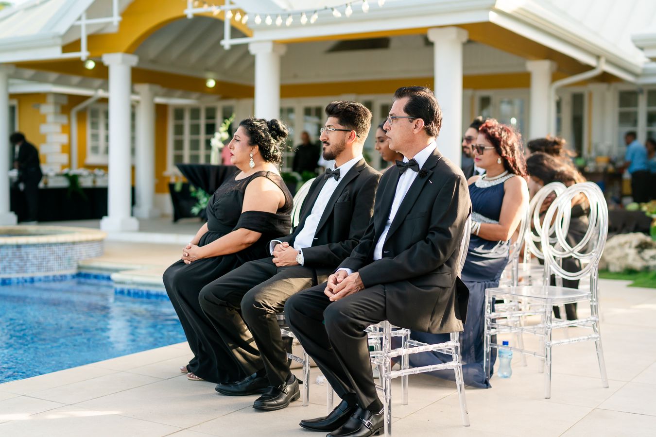 Wedding guests seated by pool watching ceremony in formal black and navy attire.