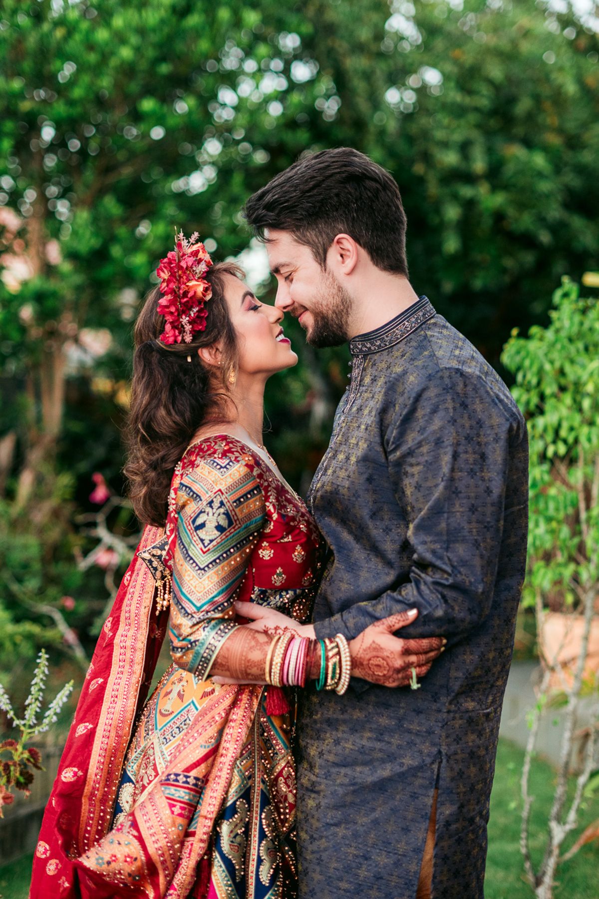 Indian couple in traditional wedding attire sharing a romantic moment outdoors, bride in red saree with floral hair accessory.