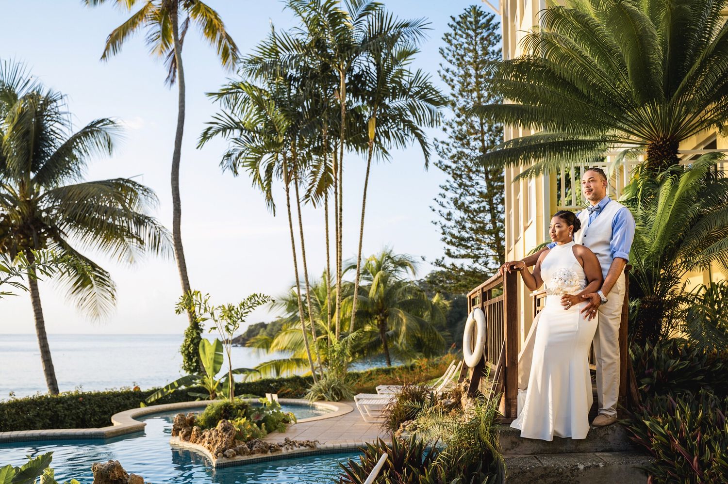 Couple standing on a tropical resort balcony overlooking a pool and ocean, surrounded by lush palm trees at sunset.