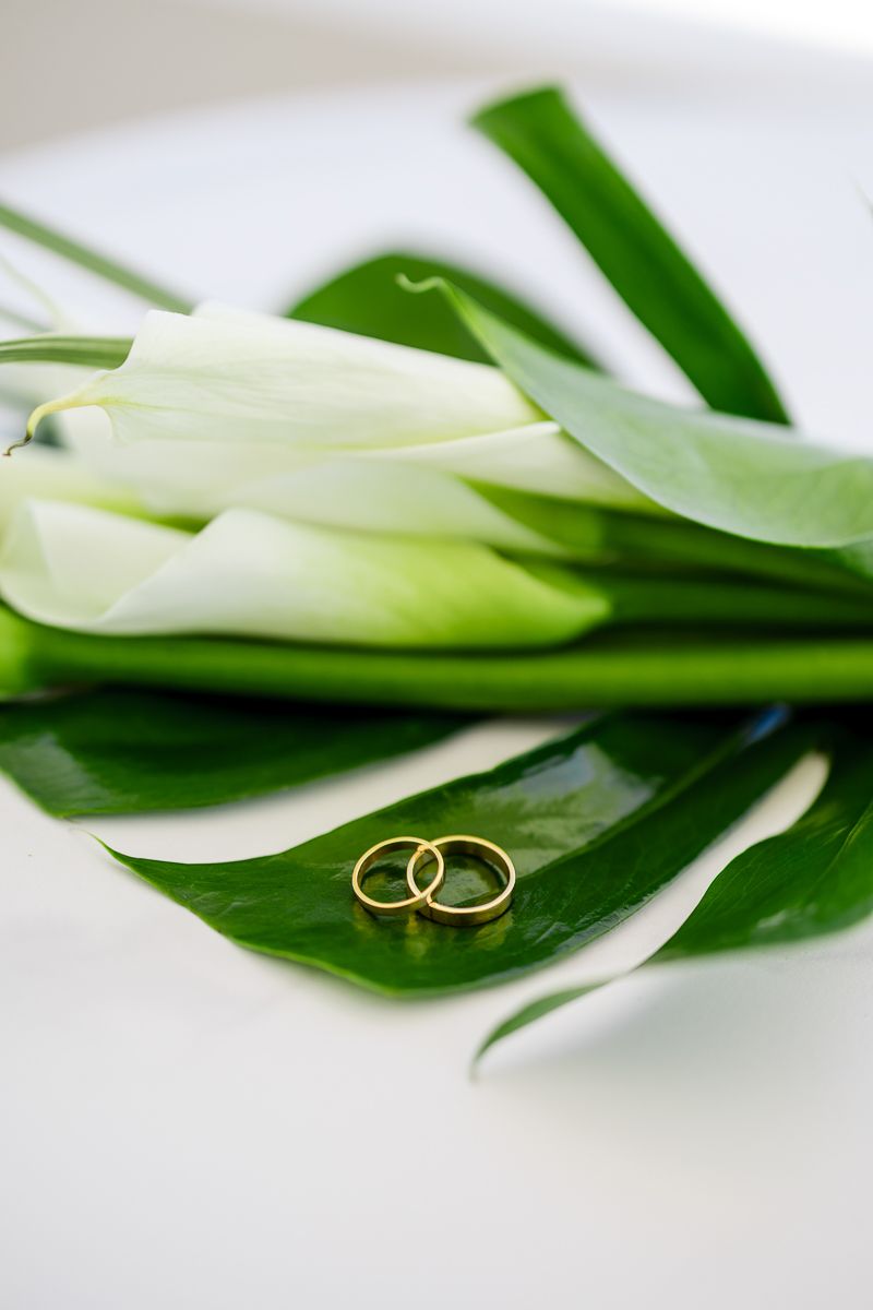 Two gold wedding rings rest on a tropical monstera leaf beside white calla lilies on a white surface.