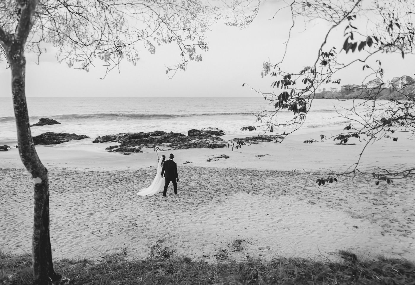 Black and white photo of bride and groom standing on a sandy beach with ocean waves behind.