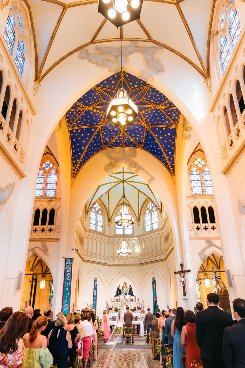 Elegant church wedding ceremony with ornate blue and gold vaulted ceiling, stained glass windows, and guests gathered in pews.