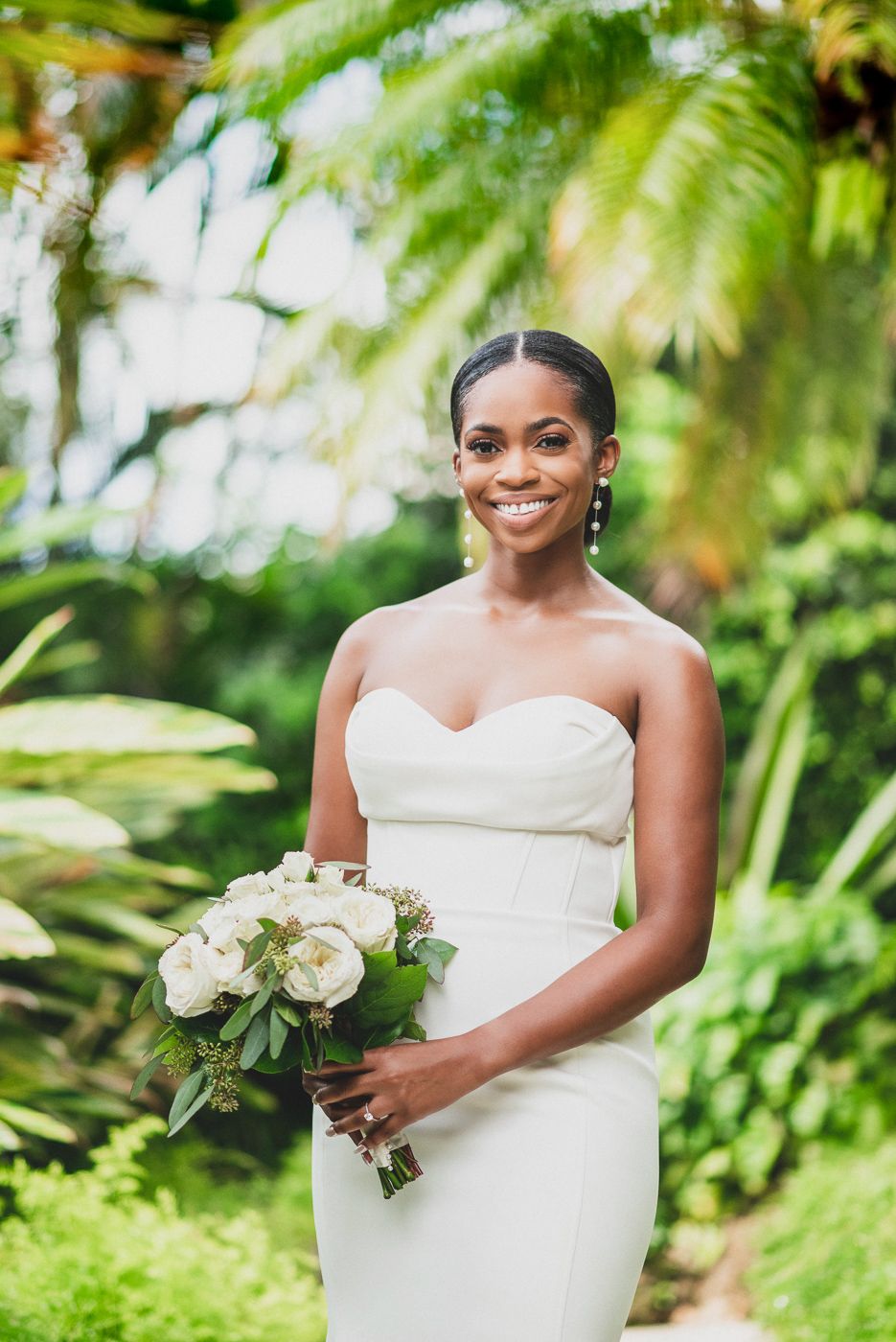 Bride holds white floral bouquet while posing against lush tropical greenery.