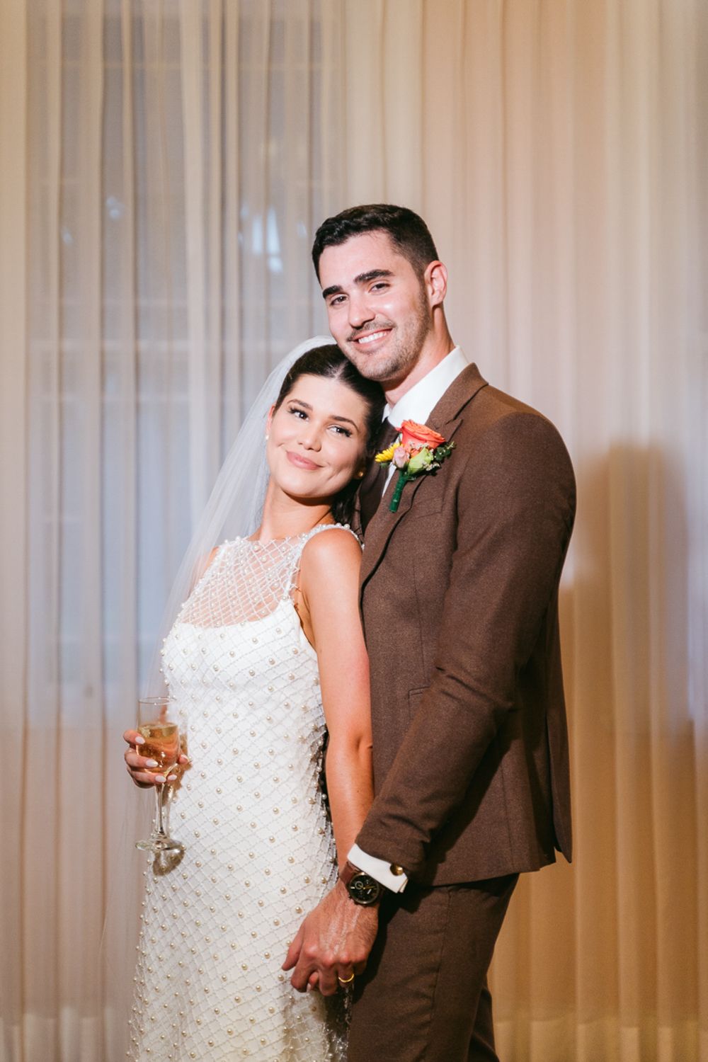 A newlywed couple poses in front of a sheer curtain backdrop, with the bride in white and groom in brown attire.