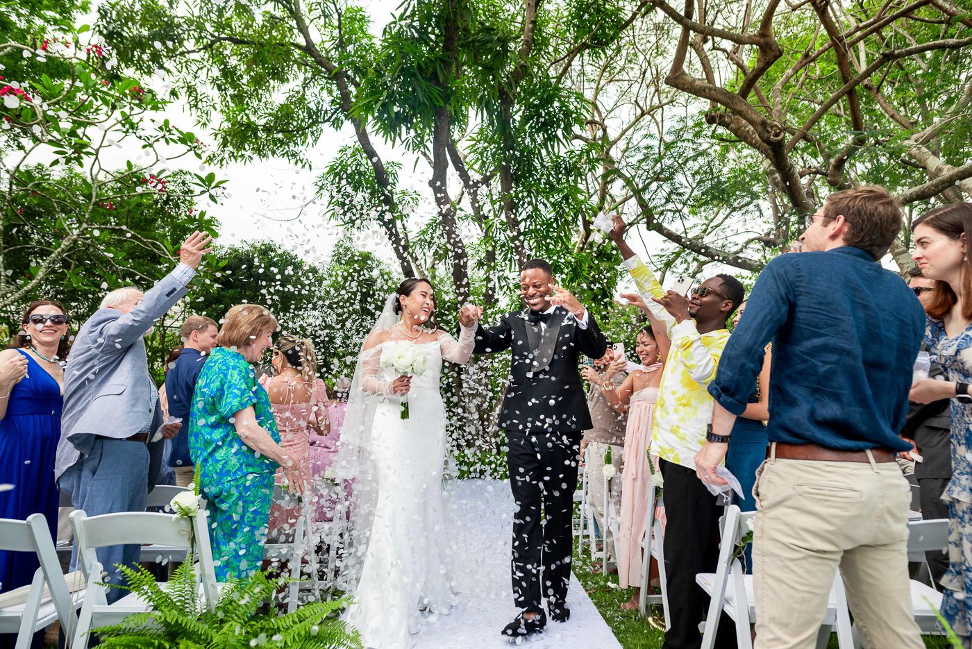 Wedding guests throw confetti as the newlyweds walk down the aisle under tropical greenery.