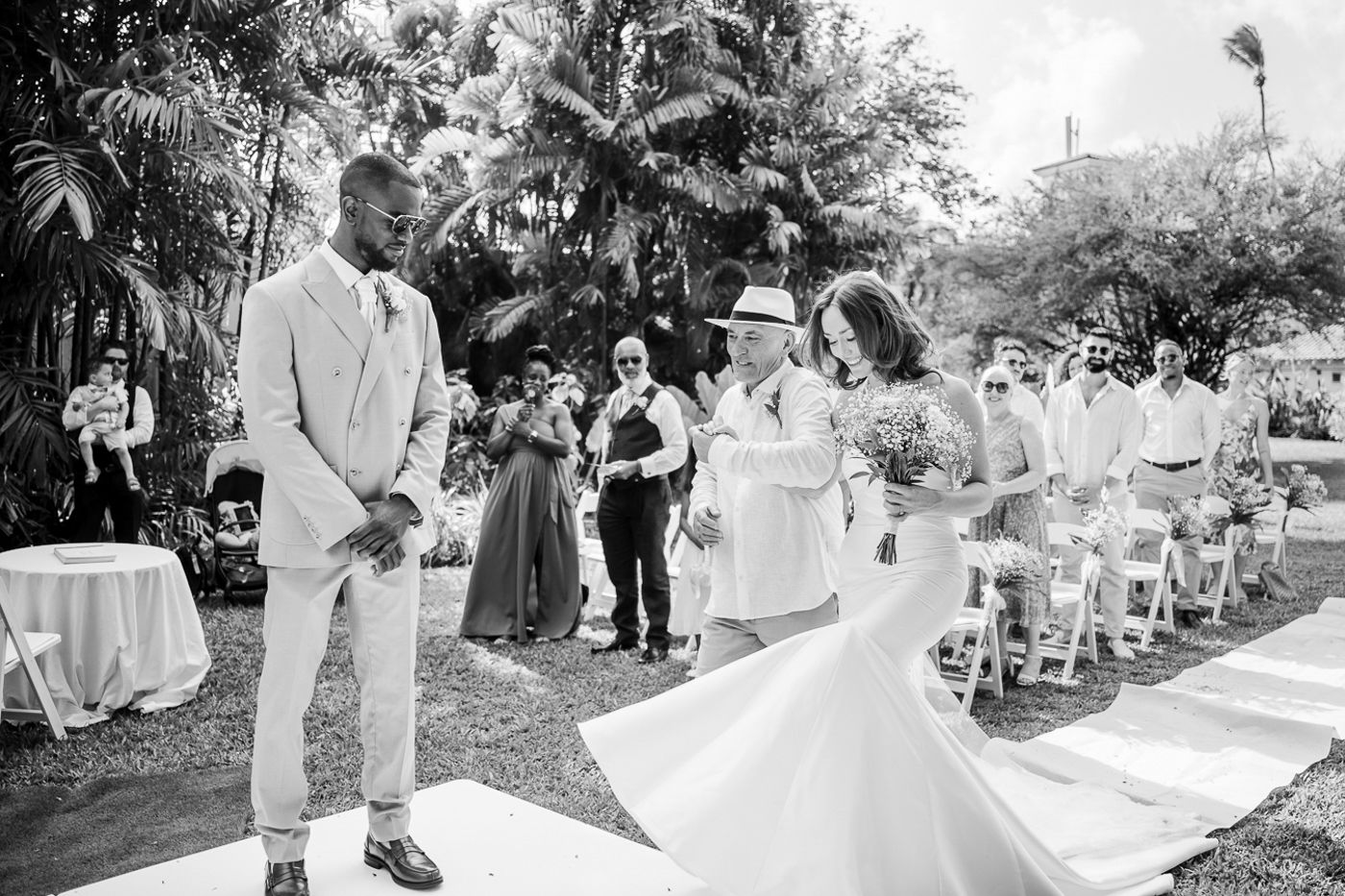 Black and white photo of a tropical garden wedding ceremony with guests seated on white chairs.
