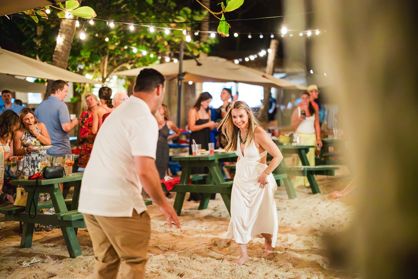 Couple dancing barefoot on sandy beach bar with string lights and green picnic tables at tropical night party.
