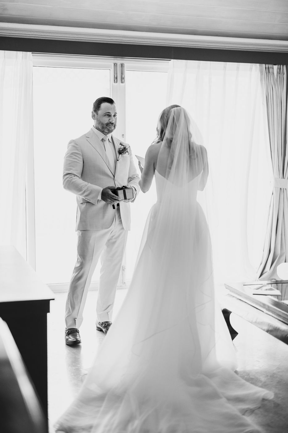 Black and white photo of a groom presenting a gift to his bride in a wedding dress and veil before their ceremony.