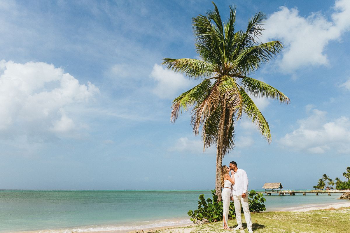 Couple embracing under a palm tree on a tropical beach with turquoise water and blue sky.
