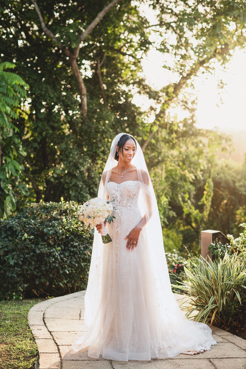 Bride in a strapless lace wedding gown with veil holds white bouquet, standing on stone path in sunlit garden.