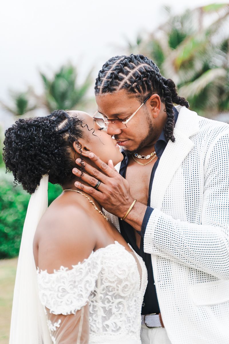A couple shares a romantic kiss outdoors, dressed in wedding attire with palm trees in the background.