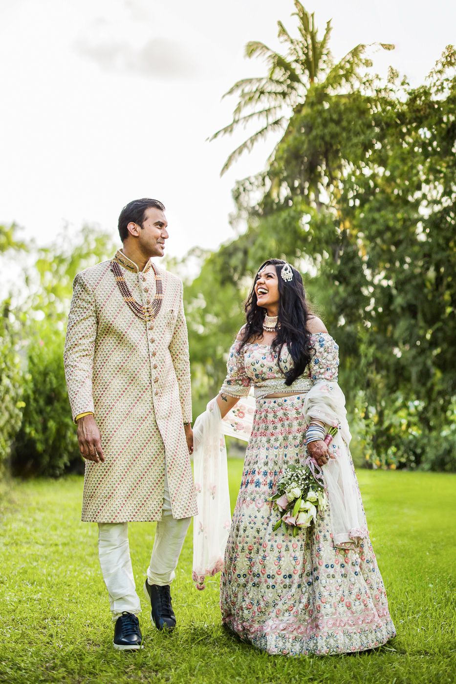 Indian couple in traditional wedding attire walking on green lawn, groom in sherwani, bride in floral lehenga.