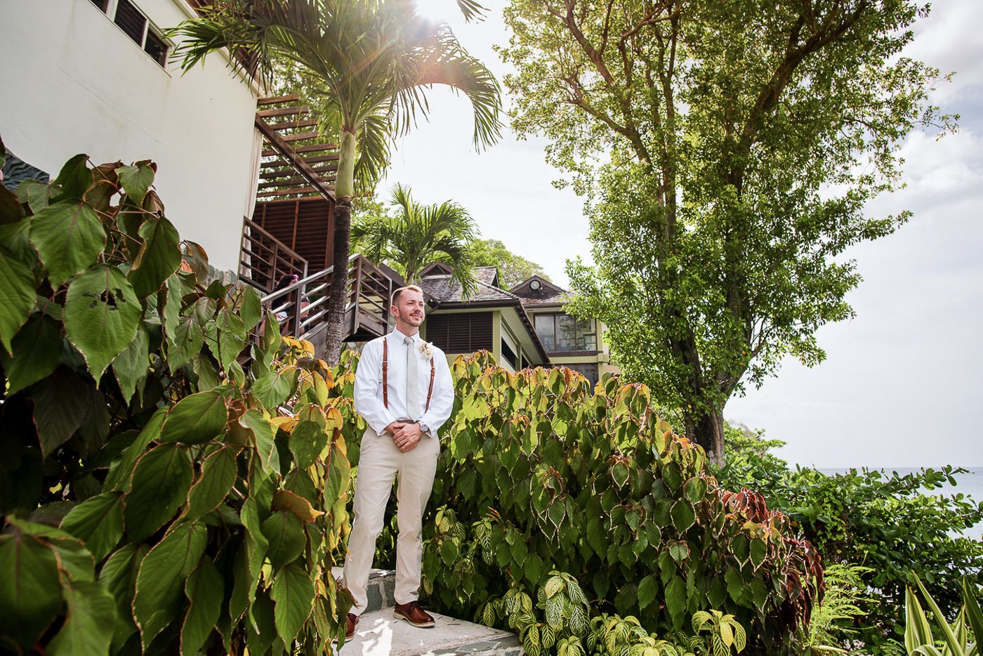 Tropical garden setting with groom in light colored suit and suspenders on stone path.