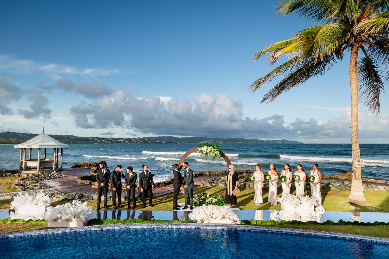 Seaside wedding ceremony setup with palm trees, ocean views, and a wooden gazebo on a tropical beach.