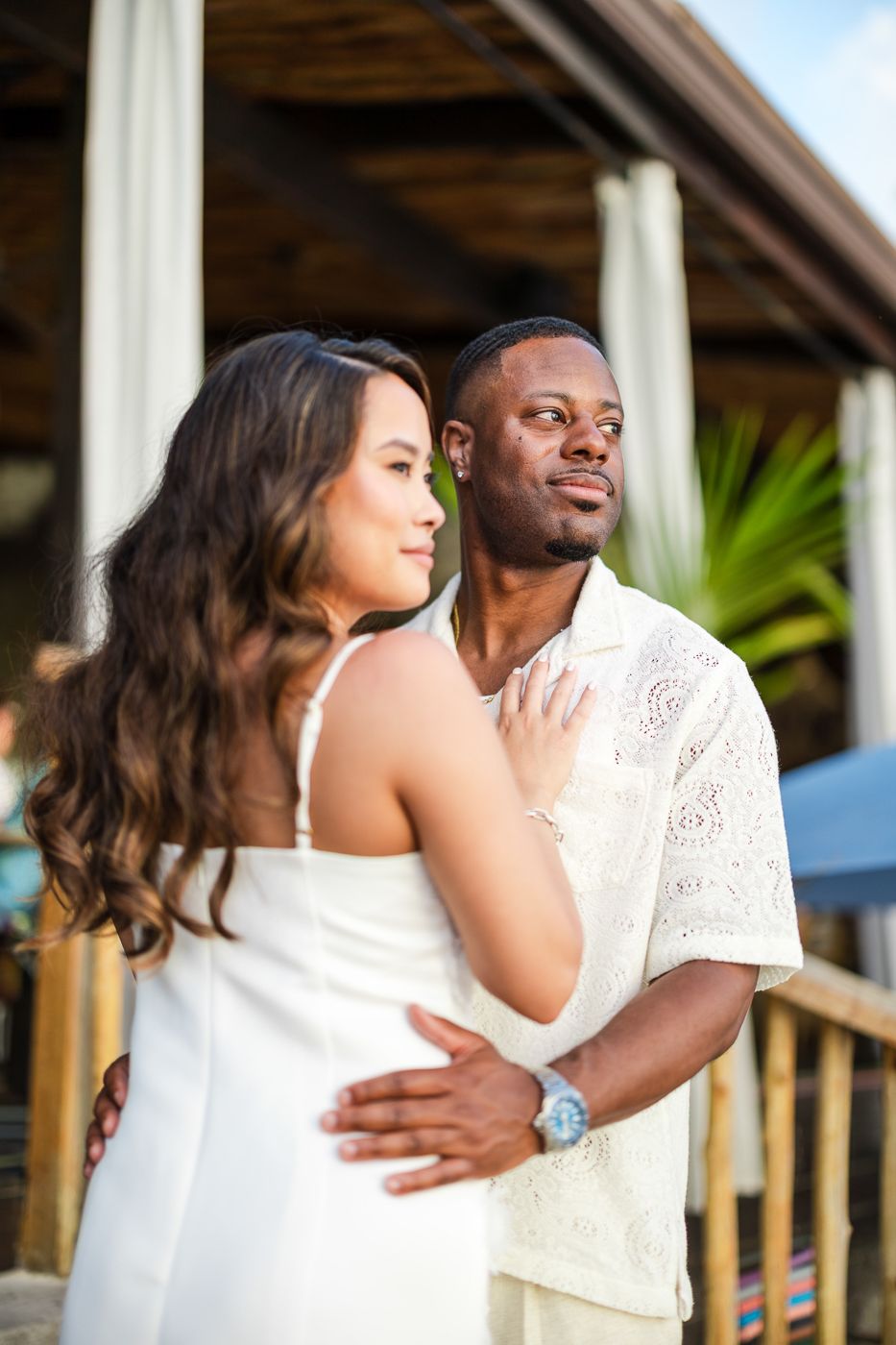 A couple in white outfits share a tender moment together on a tropical patio.