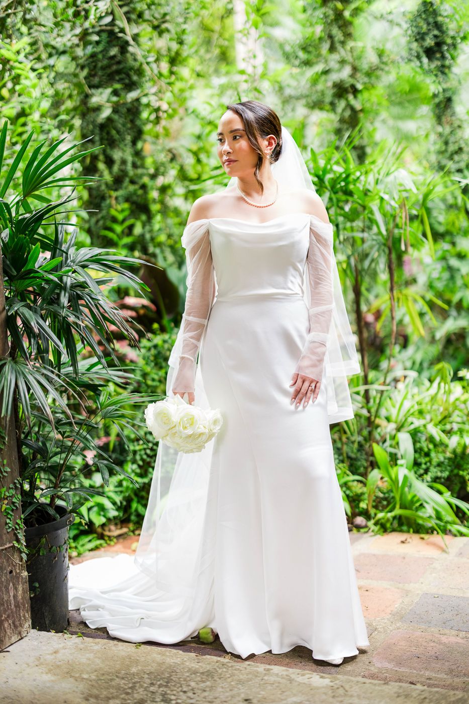Bride in flowing white dress poses gracefully in a tropical garden setting.