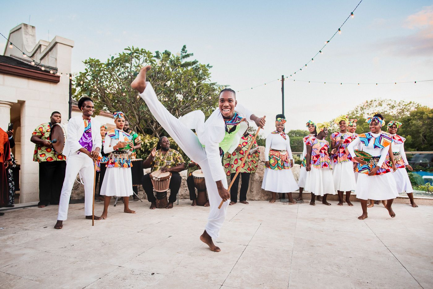 A capoeira performer in white clothing demonstrates a high kick during a lively outdoor cultural celebration.