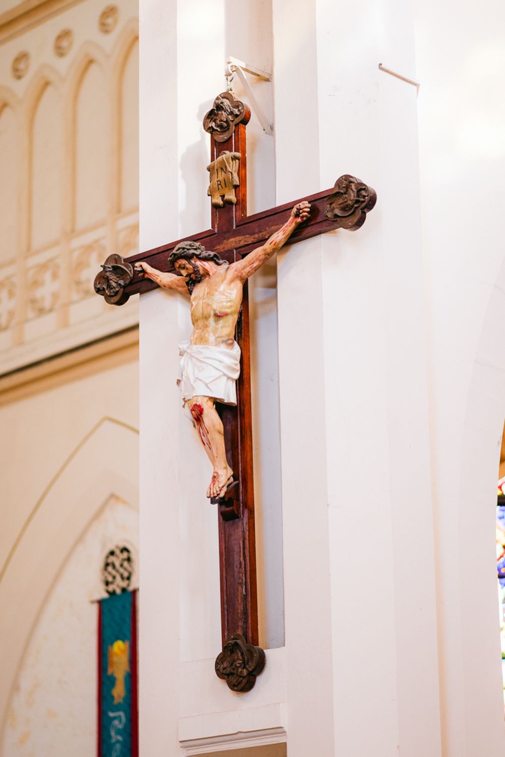 Religious crucifix mounted on white church wall with ornate architectural details in background.