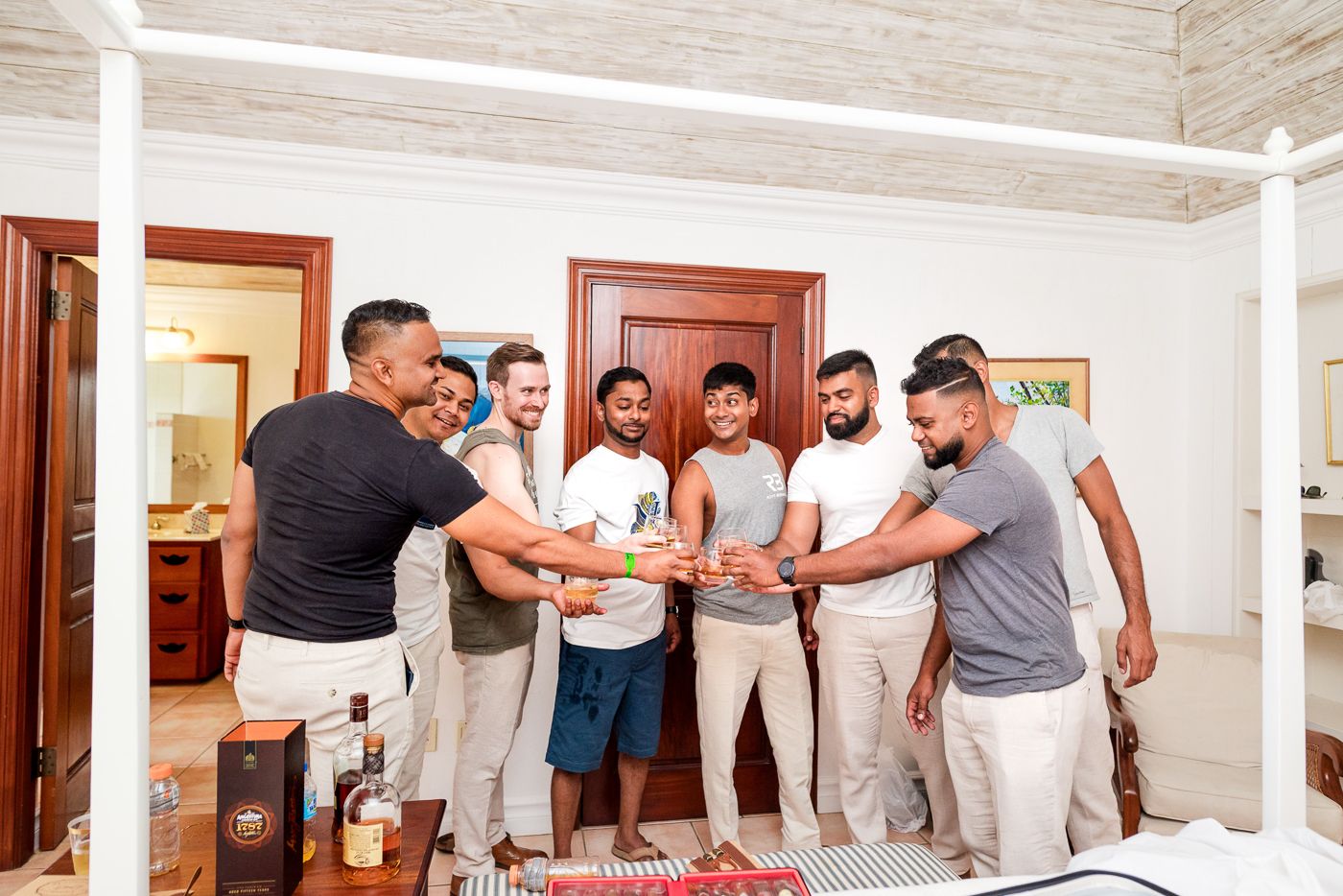 Groomsmen in casual attire gathering for a pre-wedding toast in white interior room.