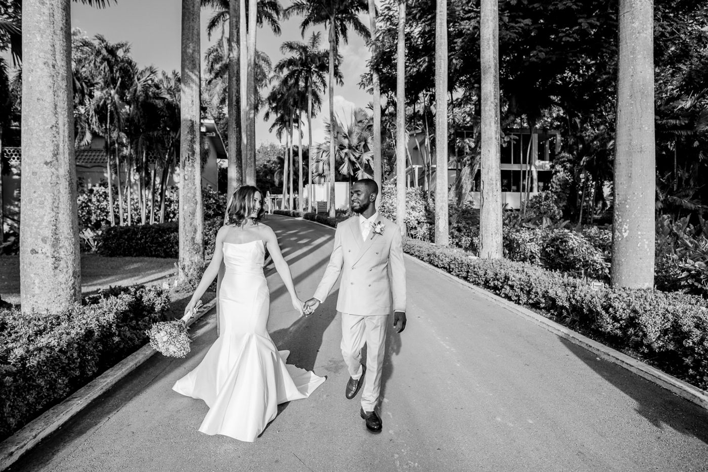 A couple walks hand in hand down a palm tree lined pathway in black and white.