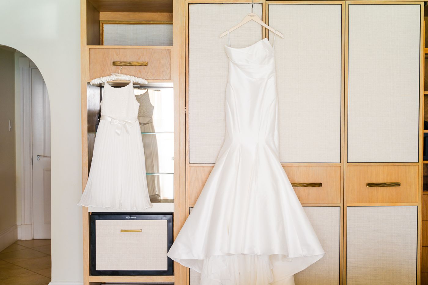 Classic white wedding dresses displayed on hangers against light wood and white walls.