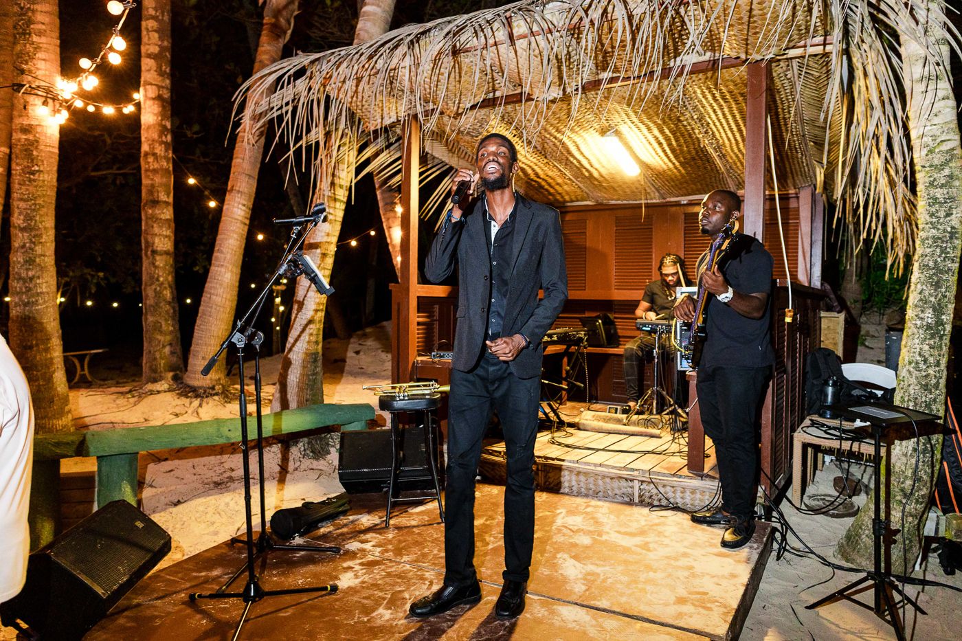 Live band performing outdoors at night under string lights and palm thatched roof at a tropical venue.