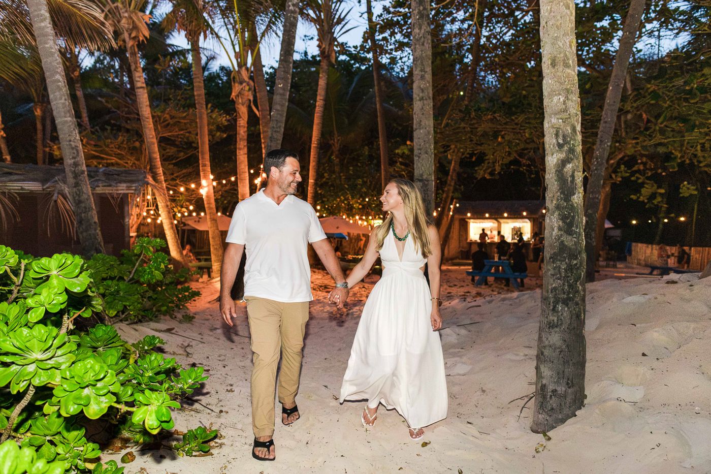 Couple holding hands walking on sandy beach path at dusk surrounded by palm trees and string lights.