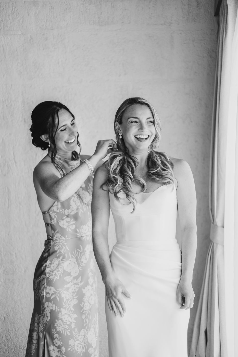 Black and white photo of a bride laughing as her bridesmaid helps fasten her earring before the wedding ceremony.