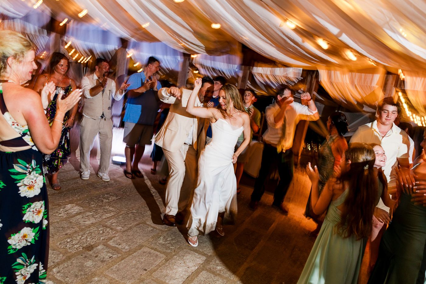 Bride and groom dancing joyfully with wedding guests under draped fabric ceiling at lively reception.