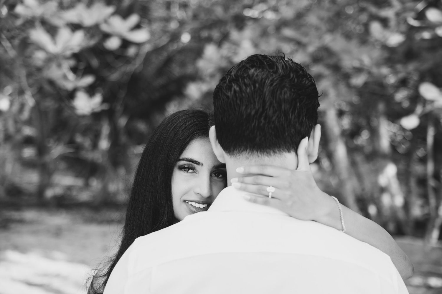 Black and white photo of a couple embracing outdoors, woman smiling over mans shoulder, trees in background.