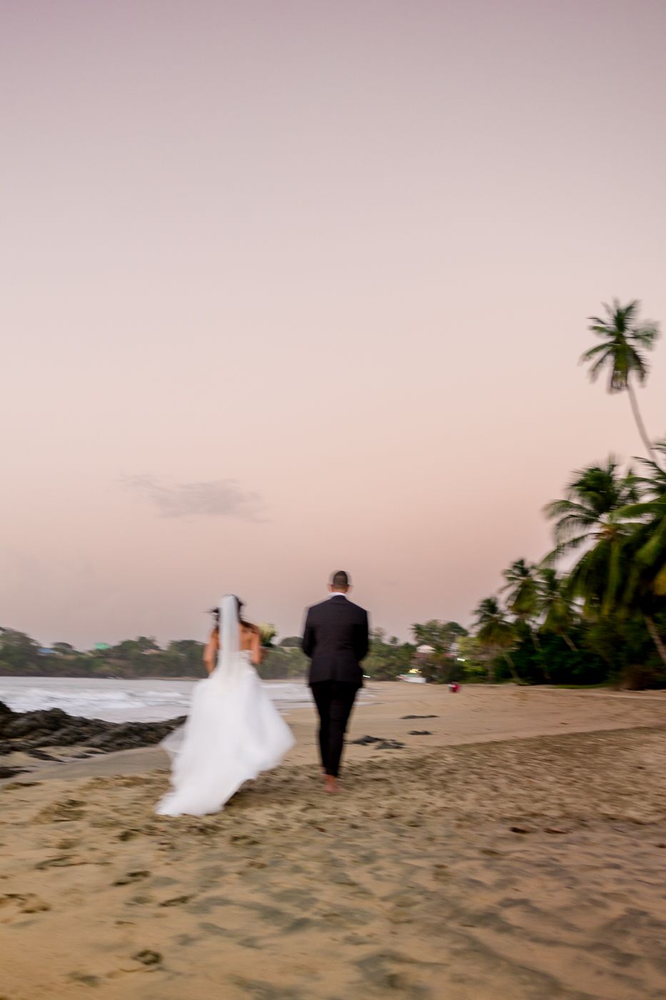 Bride and groom walking hand in hand along a tropical beach at sunset with palm trees.