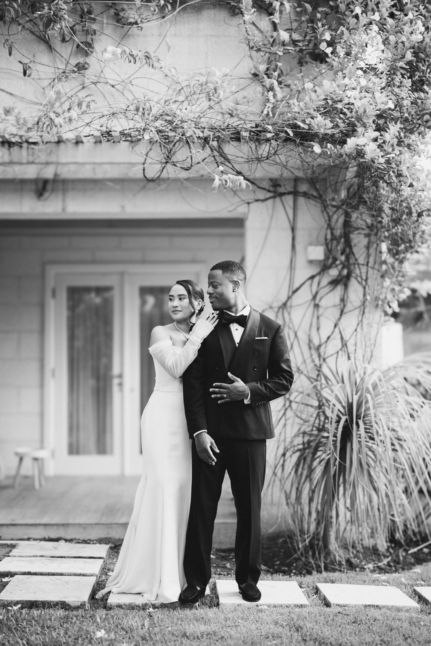 Black and white photo of a bride and groom embracing outdoors near a building with climbing vines on their wedding day.