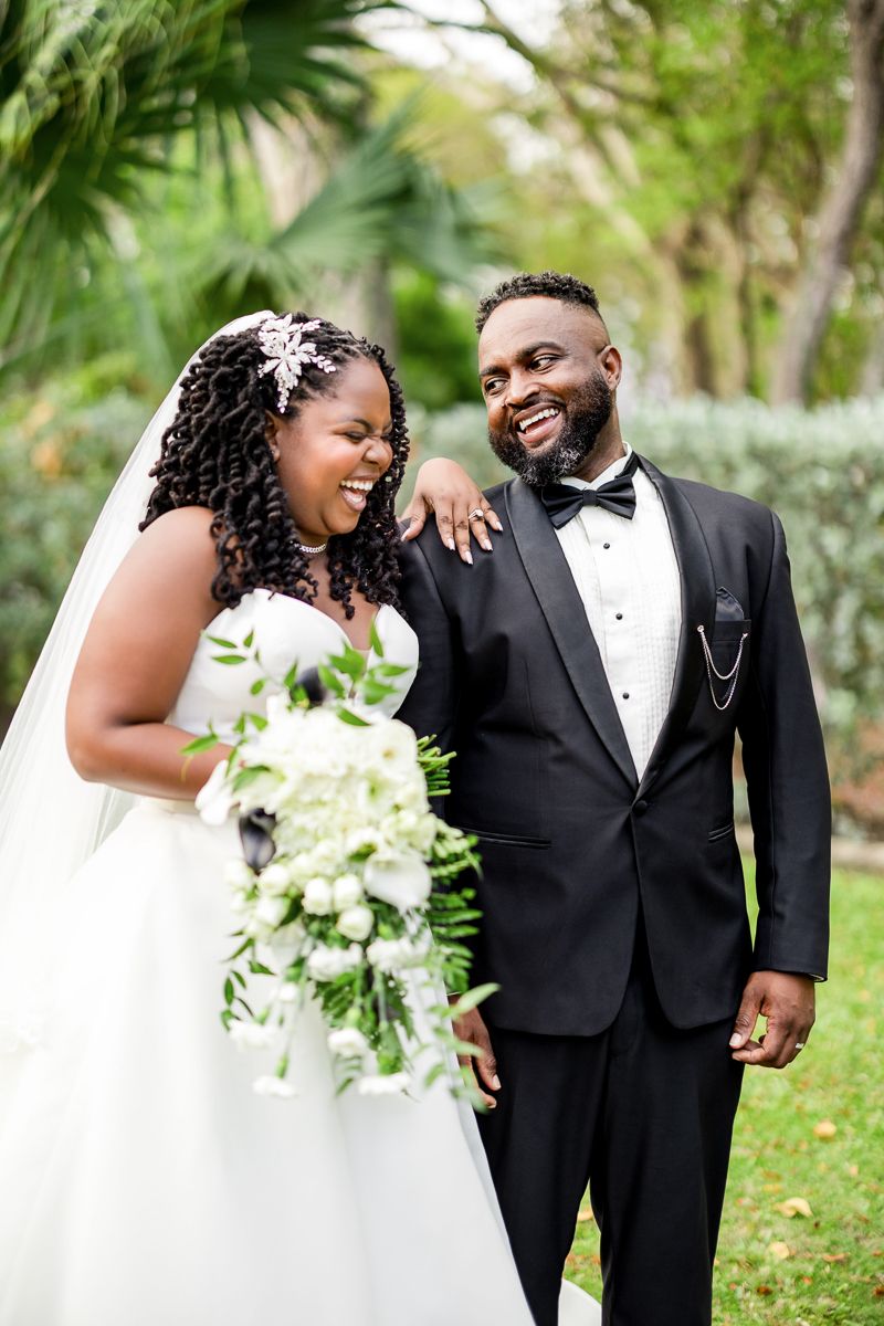 Joyful bride and groom in black tuxedo laughing together outdoors, bride holding white floral bouquet on wedding day.