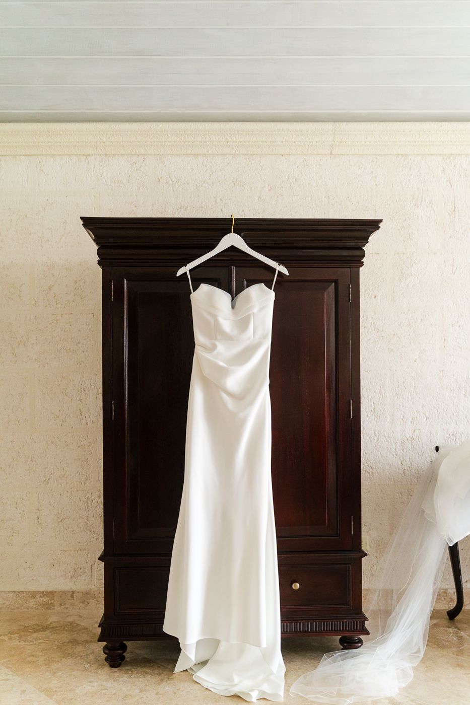 Elegant white satin strapless wedding gown hanging on a dark wood armoire with a flowing veil in a luxury bridal suite.