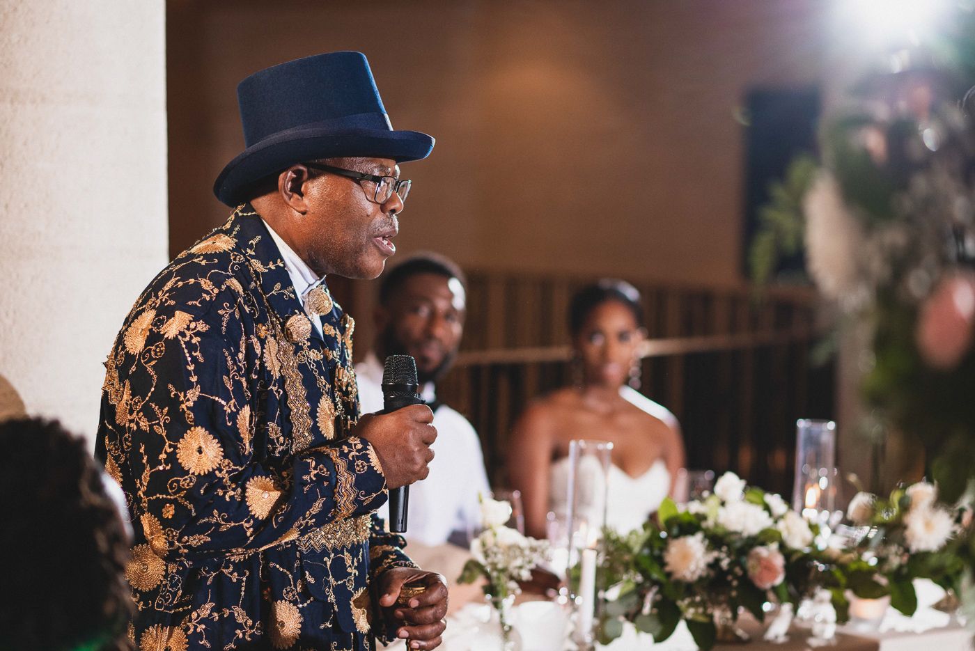 Wedding reception guests listening to speeches at tables decorated with floral centerpieces.