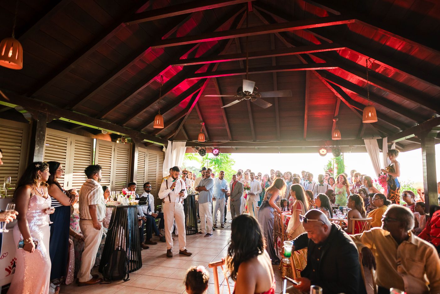 Wedding reception venue with high wooden beams and red ambient lighting filled with celebrating guests.