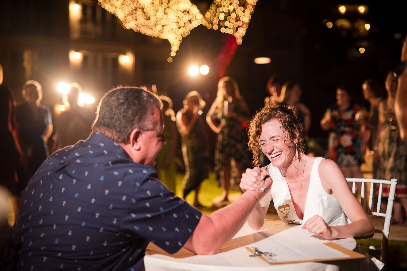 Two people engaged in an arm wrestling match at a lively nighttime reception celebration.