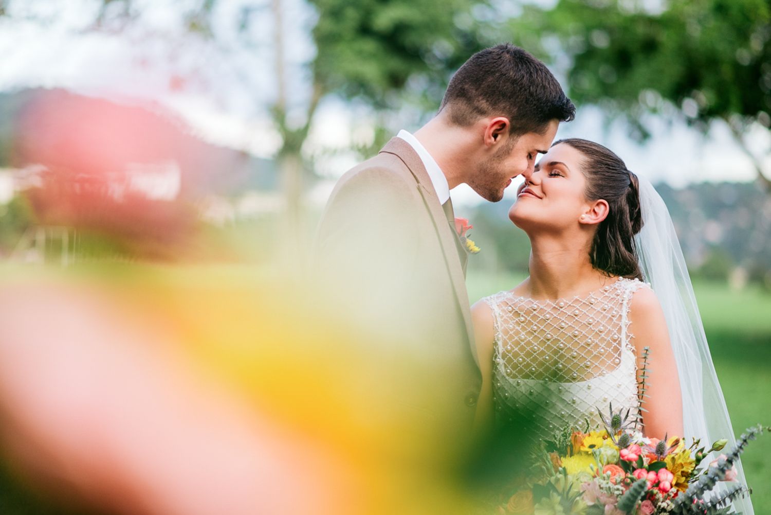 Romantic wedding couple shares tender kiss in a summer garden with sunlit bokeh and vibrant flowers.