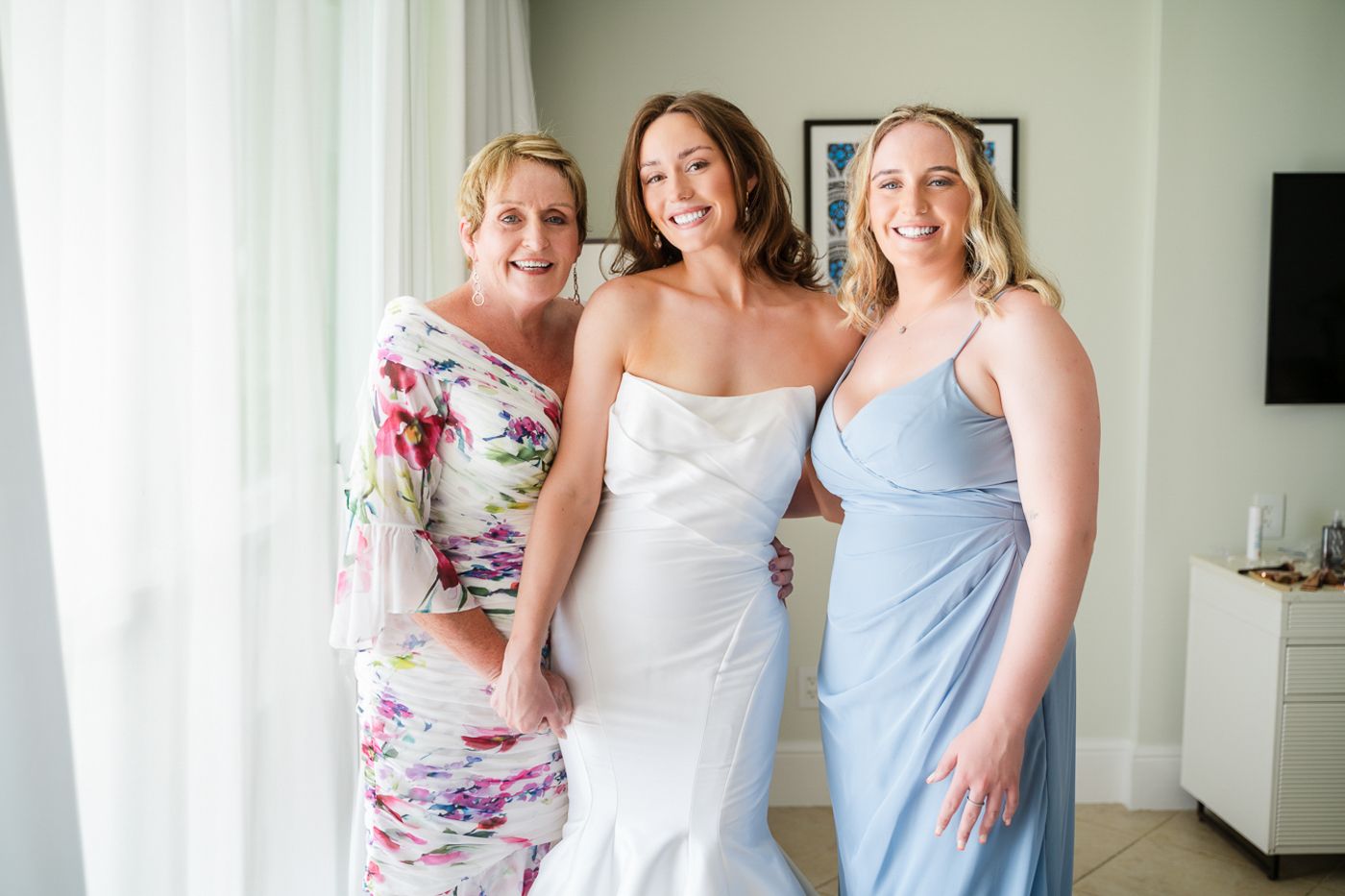 Three women pose together, wearing elegant dresses in floral and pastel colors for a wedding celebration.