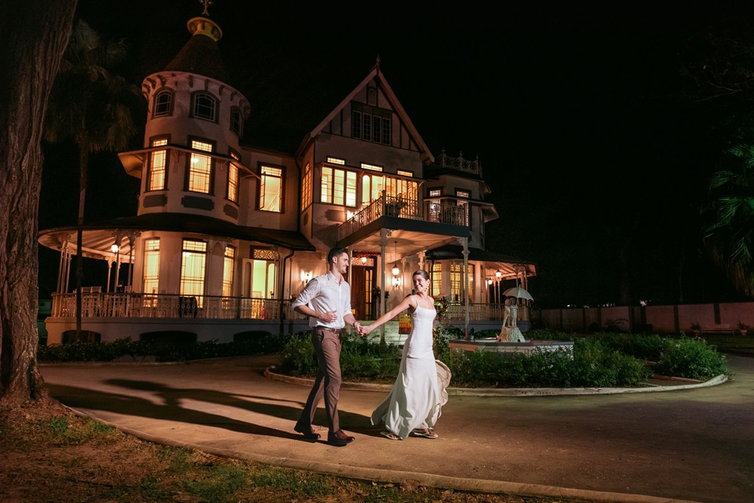 A wedding couple poses for a candid nighttime photo session in front of an illuminated historic mansion.