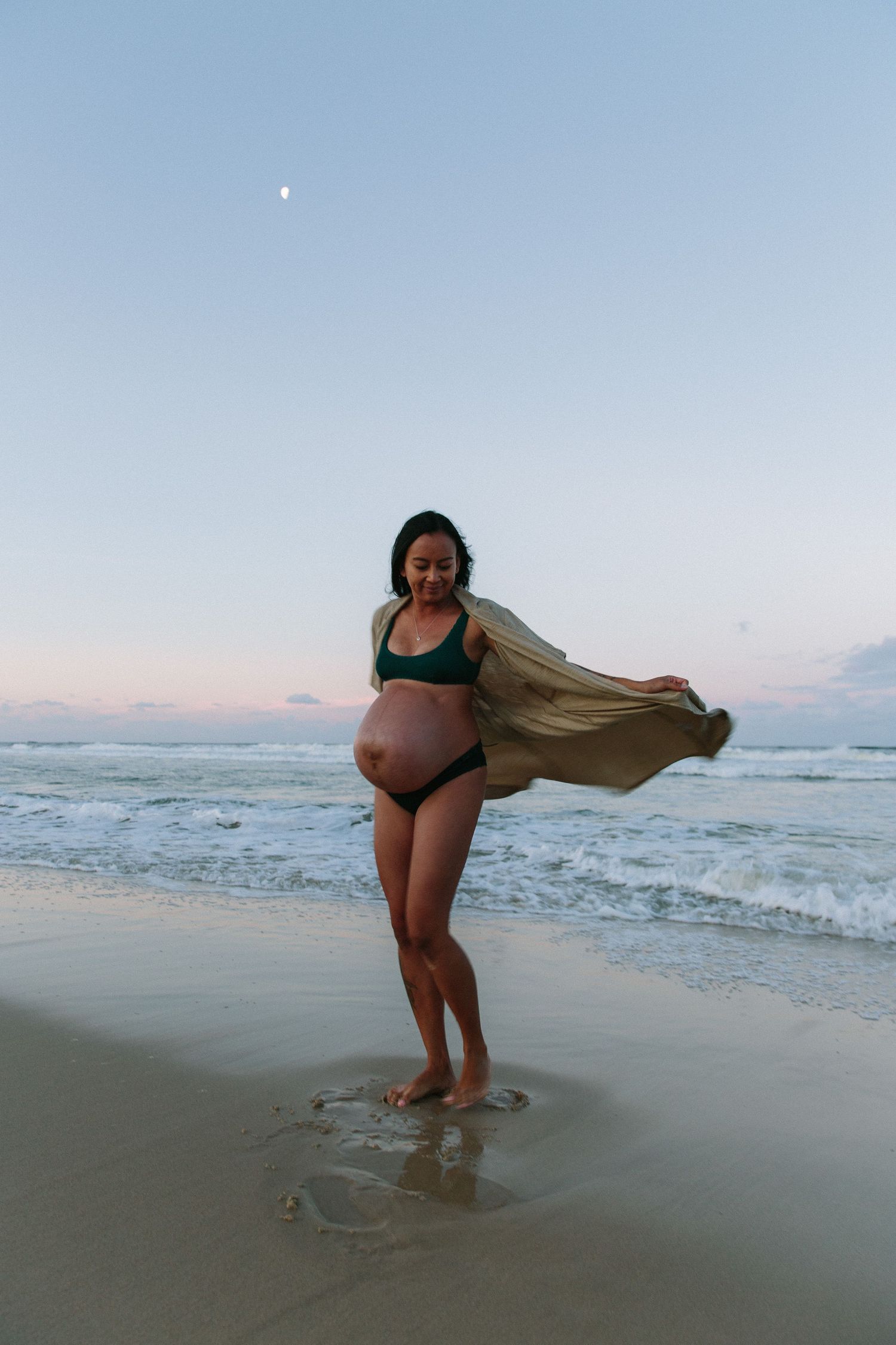 A person in black swimwear poses with a flowing fabric on a beach at sunset.