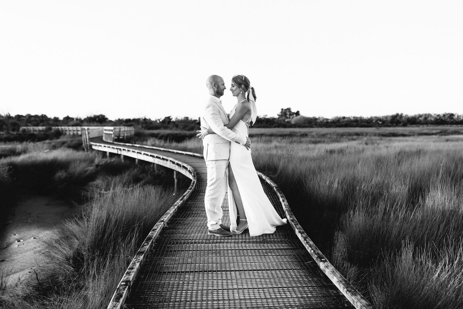 Wooden boardwalk path extends through tall marsh grasses in black and white landscape photography.