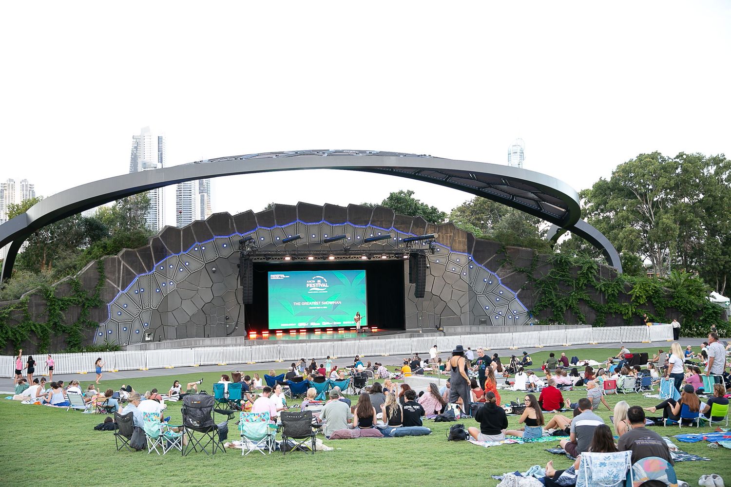 Panoramic view of outdoor event with crowd gathered on lawn in front of stage under bridge.