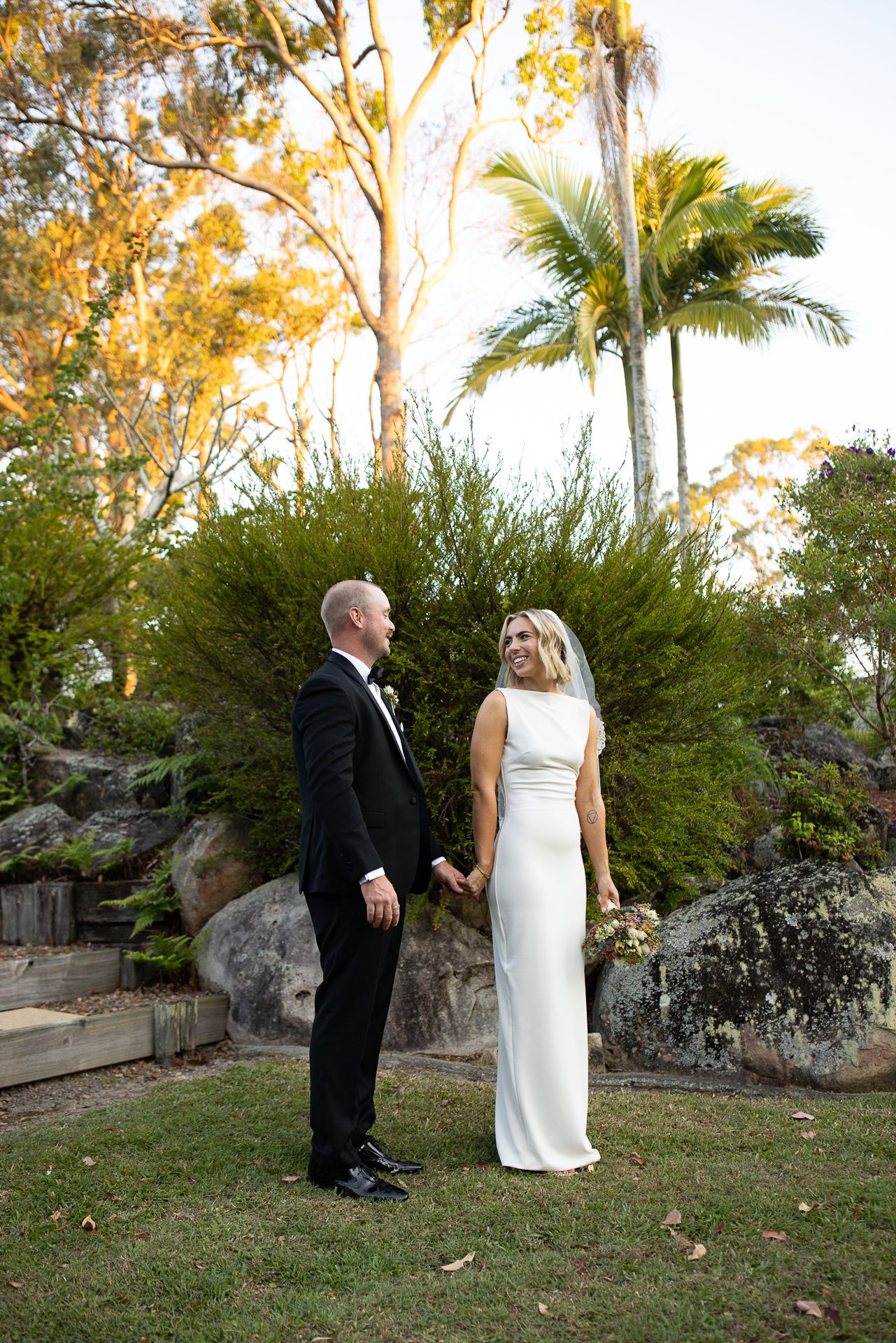 A couple in formal wedding attire walks through a garden setting with palm trees at sunset.