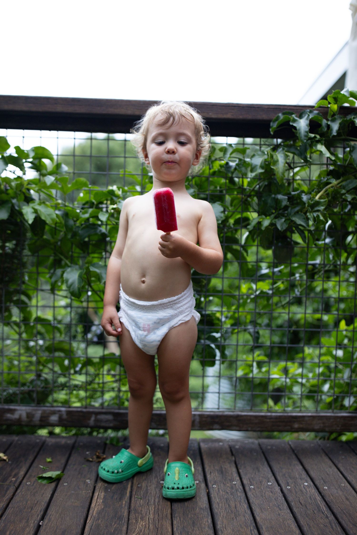 Small child in diaper and green shoes standing on wooden deck with vines in background.